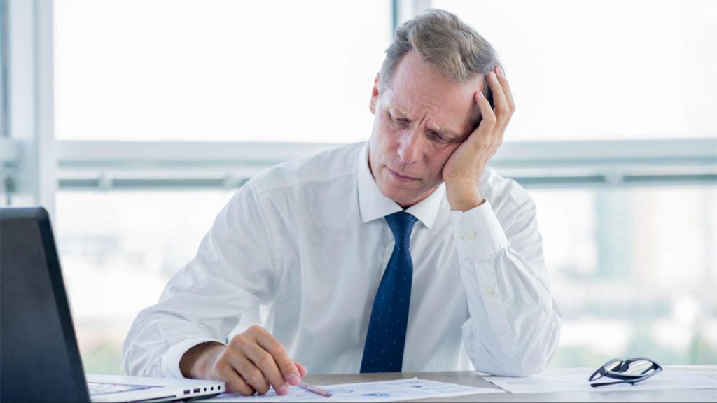Businessman in a shirt and tie holding his head with one hand while seated at a desk Caption: Chronic stress and emotional strain can build silently in high-demand professions, affecting performance and judgment.