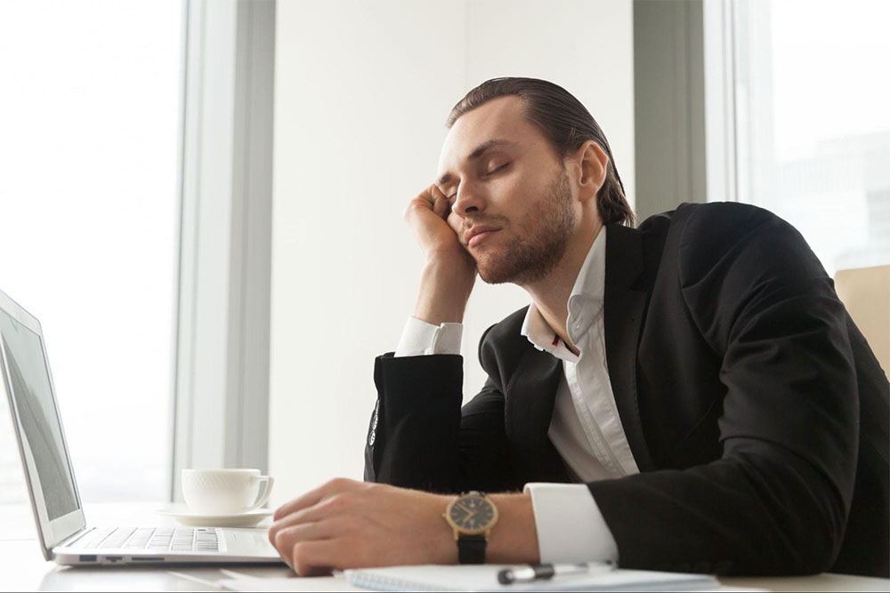 A man in formal attire resting his head on his hand while looking at a laptop screen.