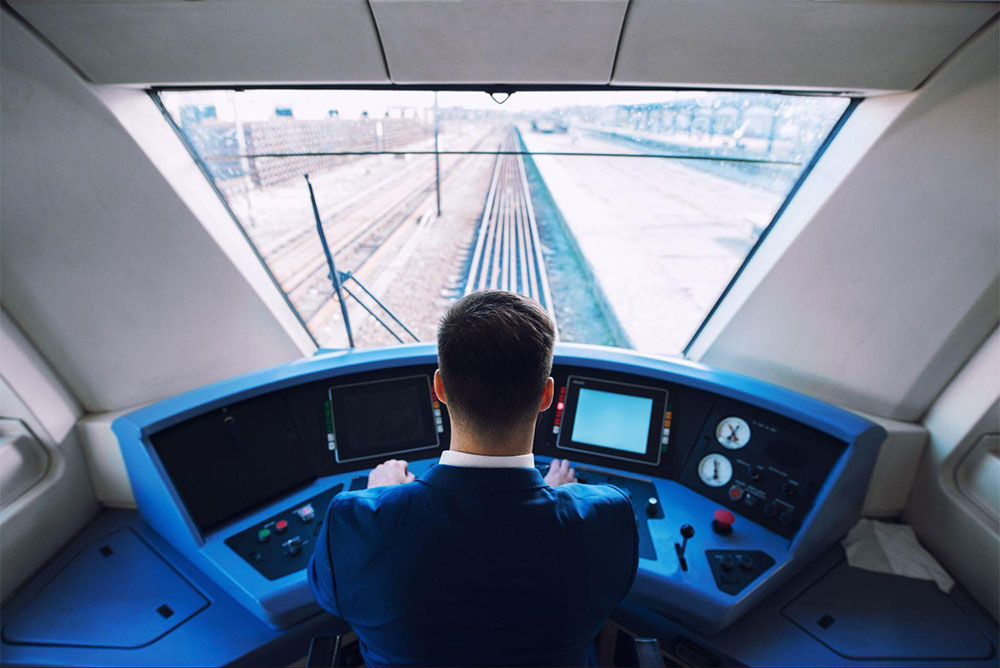 Rear view of a train operator seated inside a control cabin overlooking train tracks.